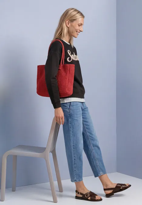 Sweatshirt met lange mouwen en Artwork black coffee Junge Frau trägt rote Tasche, Jeans und Pullover, steht lächelnd neben einem Stuhl vor hellem Hintergrund.