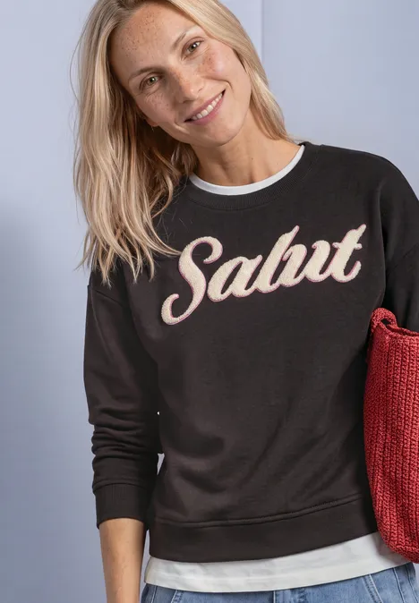 Sweatshirt met lange mouwen en Artwork black coffee Eine lächelnde blonde Frau trägt ein schwarzes Sweatshirt mit dem Schriftzug "Salut" und hält eine rote Tasche.