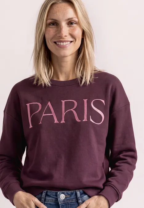 Frau lächelt und trägt einen weinroten Pullover mit der Aufschrift 'PARIS'.