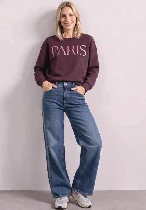 Junge Frau trägt lila Sweatshirt mit Paris-Schriftzug, weite Jeans und Sneaker, steht lächelnd vor heller Wand.