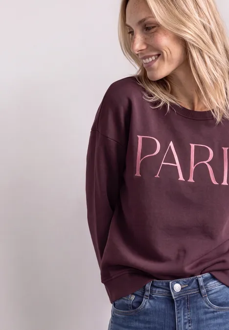 Lächelnde Frau in Jeans und weinrotem Pullover mit Aufschrift 'PARIS' vor schlichtem Hintergrund.