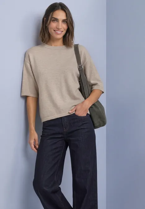 Vrouw lacht in beige kort trui, donkere wijdere jeans en groene schoudertas voor lichte achtergrond.
