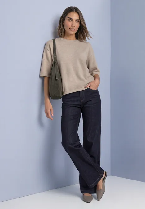 Een vrouw draagt een beige trui, donkere jeans en grijze schoenen voor een lichte achtergrond.
