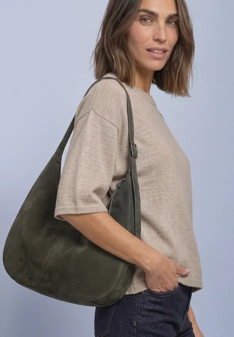 Vrouw in beige shirt en donkere jeans draagt een grote groene suède schoudertas en glimlacht subtiel.