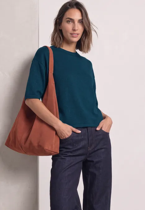 Een vrouw draagt een blauwgroen shirt, een donkere spijkerbroek en een oranje tas over haar schouder.
