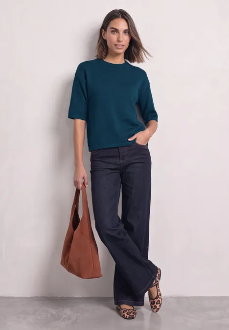 Een vrouw draagt een petrolgroene top, donkerblauwe wijde jeans, luipaardprint ballerina’s en houdt een bruine tas vast.