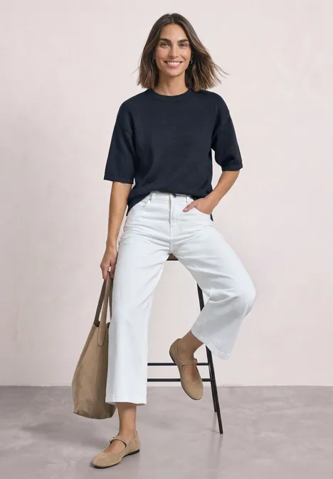 Vrouw in zwart shirt, witte wijde broek, beige loafers en tas, glimlachend leunend tegen een stoel.