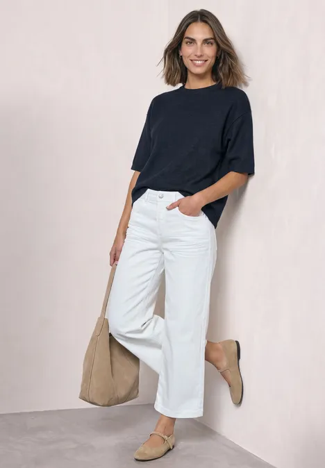 Een lachende vrouw leunt tegen een lichte muur, met een marineblauw shirt, witte broek, beige tas en bijpassende schoenen.