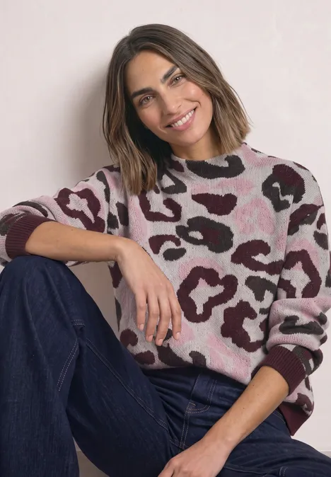 Une femme souriante est assise, portant un pull rose à motifs bruns et un jean bleu.