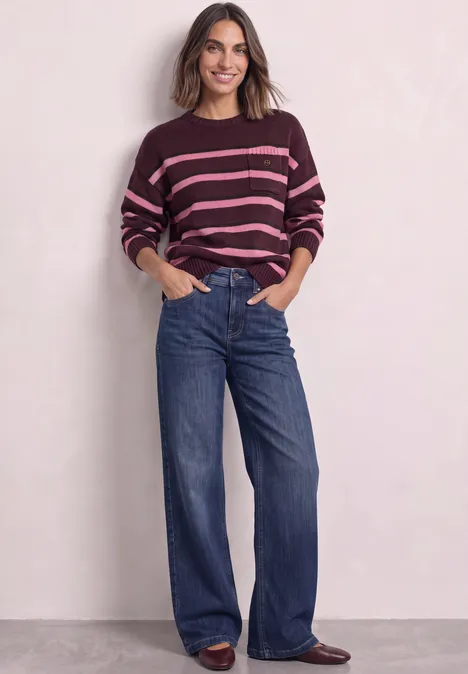 Frau mit gestreiftem Pullover und weiten Jeans vor hellem Hintergrund.