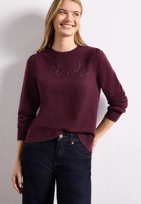 Pull avec inscription brodée rustic rouge Fröhliche Frau trägt beerenfarbenen Pullover mit aufgesticktem Schriftzug und dunkle Jeans, Hände in den Taschen.