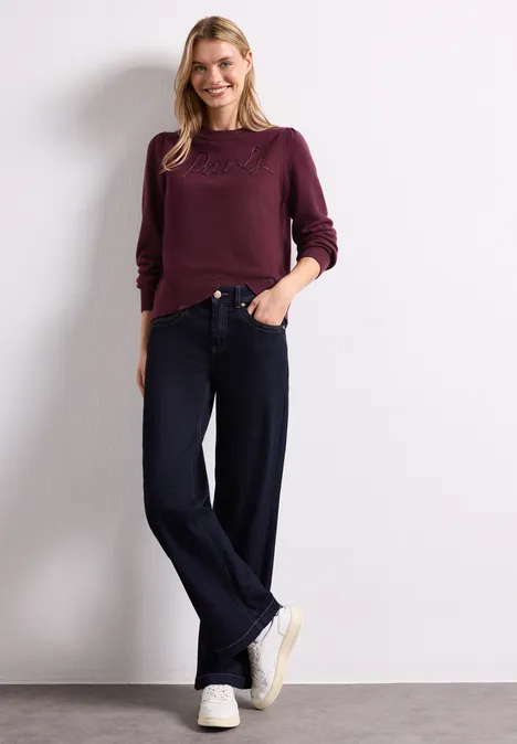 Pull avec inscription brodée rustic rouge Eine Frau trägt einen bordeauxfarbenen Pullover, weite Jeans und weiße Sneaker vor weißem Hintergrund