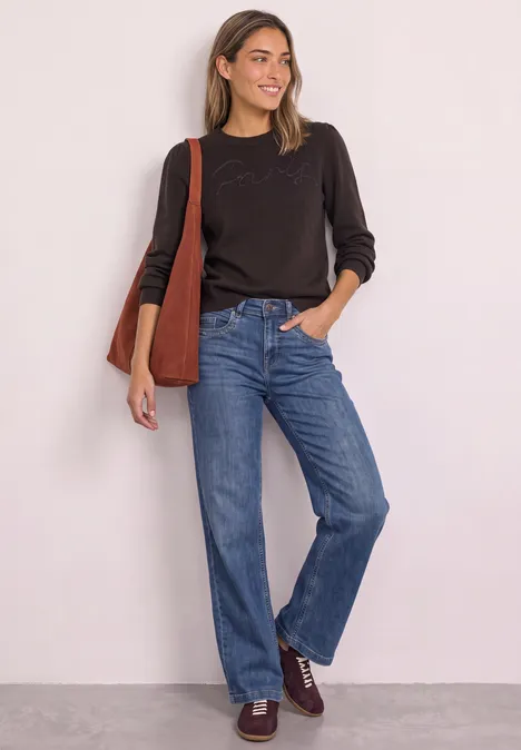 Eine Frau in dunklem Pullover mit Stofftasche trägt lässige Jeans und braune Schuhe vor heller Wand.