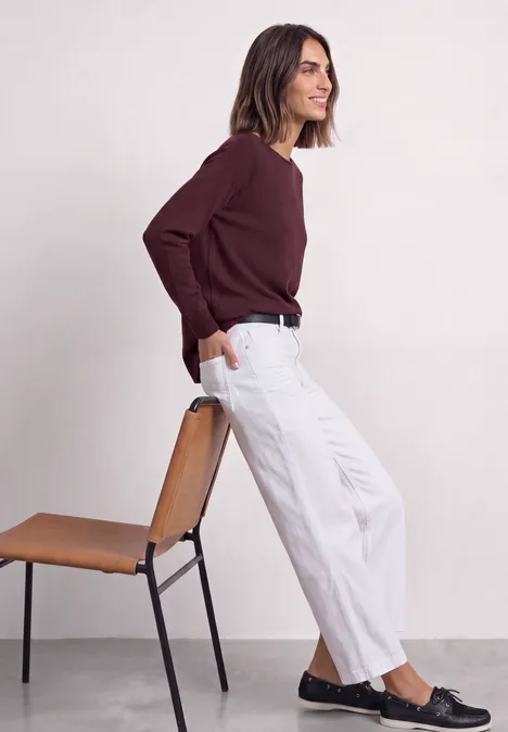 Dolman-trui met boothals rustic rouge Vrouw in witte broek en bordeaux trui, leunt ontspannen tegen een stoel.