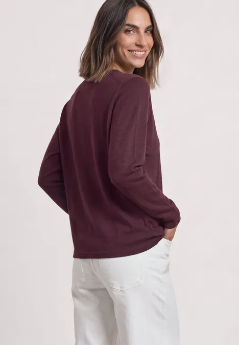 Dolman-trui met boothals rustic rouge Vrouw in bordeauxrode trui en witte broek, glimlachend opzij tegen lichte achtergrond.