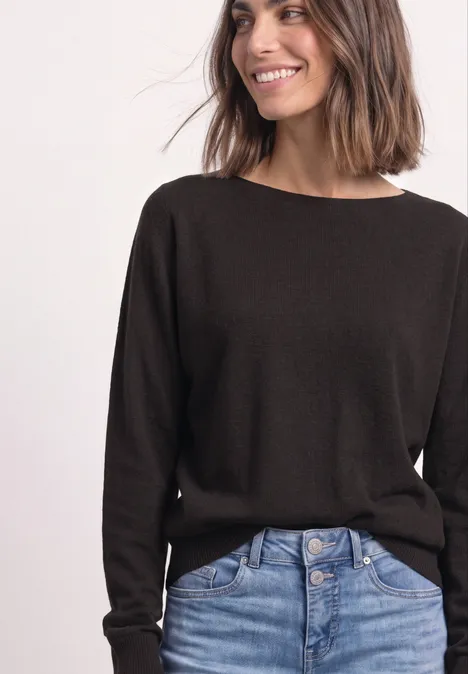 Sweter z rękawami typu dolman i dekoltem w kształcie łódki black coffee Kobieta uśmiecha się z boku, ma ciemnobrązowy sweter i dżinsy z wysokim stanem.