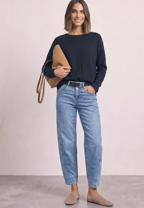Sweter z rękawami typu dolman i dekoltem w kształcie łódki darkest blue Kobieta w czarnym swetrze, niebieskich dżinsach i brązowych butach trzyma beżową kopertówkę na jasnym tle.