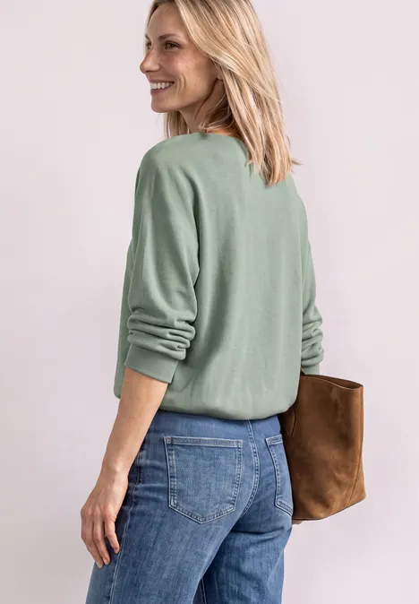 Frau in olivgrünem Pullover und Jeans hält eine braune Clutch und blickt über die Schulter.