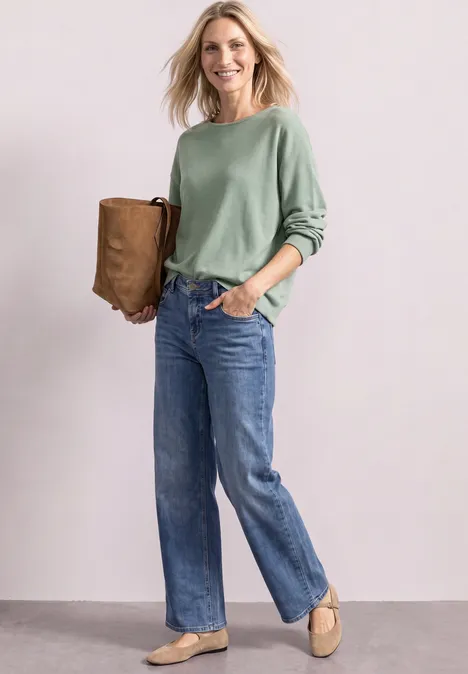 Frau in grüner Bluse und Jeans mit brauner Tasche und beigen Schuhen vor hellem Hintergrund.