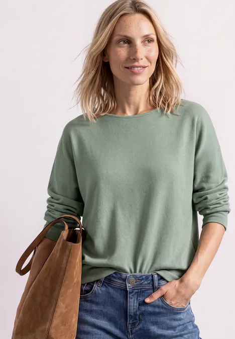 Frau in grünem Pullover und Jeans, lächelnd mit brauner Tasche vor weißem Hintergrund.