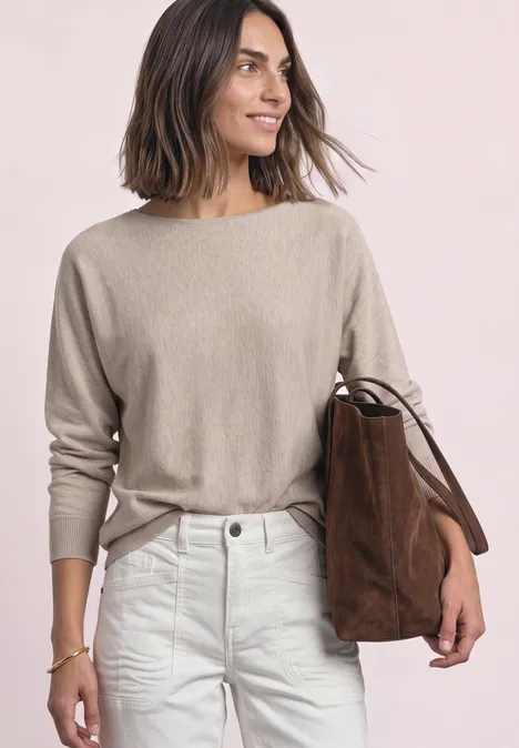 Sweter z rękawami typu dolman i dekoltem w kształcie łódki clay sand mel. Kobieta z półdługimi włosami w beżowym swetrze, białych dżinsach i brązowej zamszowej torbie.