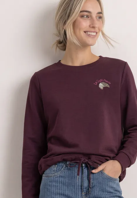 Sweatshirt met ronde hals en borduursel rustic rouge Frau in weinrotem Langarmshirt mit Tunnelzug, lächelnd vor hellem Hintergrund.
