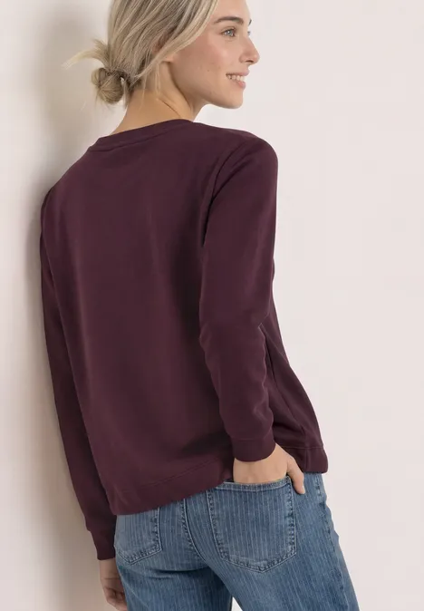 Sweatshirt met ronde hals en borduursel rustic rouge Frau in bordeauxrotem Pullover und Jeans lehnt seitlich an einer Wand.