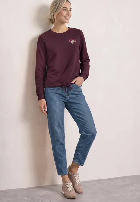 Sweatshirt met ronde hals en borduursel rustic rouge Frau lehnt an Wand, trägt bordeauxrotes Sweatshirt, blaue Jeans und bequeme Sneakers.