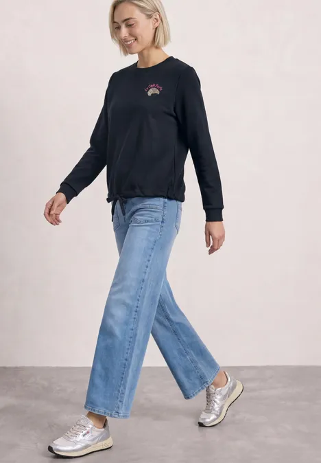 Sweatshirt met ronde hals en borduursel darkest blue Frau in schwarzem Langarmshirt, weitem Jeanshose und silbernen Sneakers vor hellem Hintergrund.