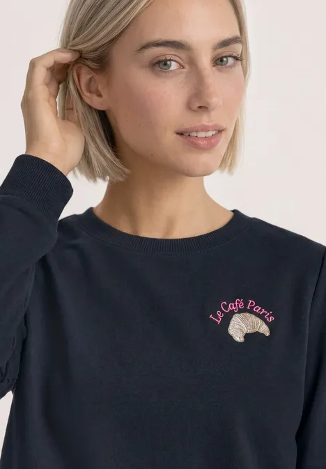 Sweatshirt met ronde hals en borduursel darkest blue Frau mit kurzem blondem Haar in dunkelblauem Pullover, bestickt mit Croissant und Schriftzug „Le Café Paris“.