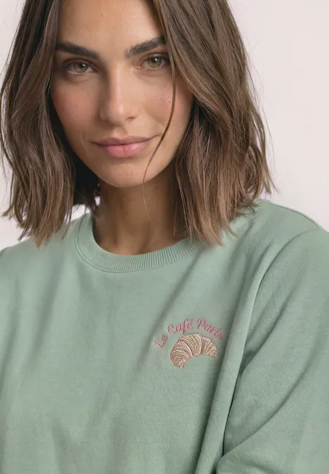 Sweatshirt met ronde hals en borduursel eucalyptus green Frau mit braunen Haaren in grünem Sweatshirt mit Croissant-Logo und Schriftzug „Le Café Paris“.