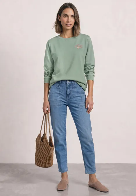 Sweatshirt met ronde hals en borduursel eucalyptus green Frau in grünem Sweatshirt, blauen Jeans, beigen Schuhen und Korbtasche vor hellem Hintergrund.