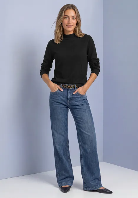 Pullover in Unifarbe mit Rundhals Black Frau mit schwarzem Pullover, weitem blauen Jeans, Gürtel und spitzen Schuhen vor neutralem Hintergrund.