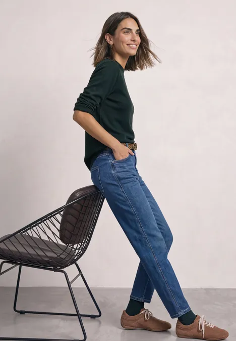 Pullover in Unifarbe mit Rundhals stormy green Eine Frau in schwarzem Pullover und blauen Jeans lehnt lächelnd an einem Stuhl vor hellgrauem Hintergrund.