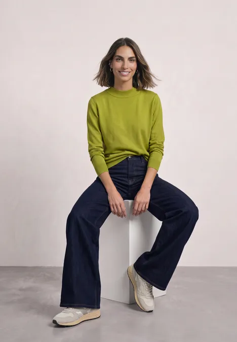 Pullover in Unifarbe mit Rundhals apple lime Eine lächelnde Frau sitzt auf einem Hocker, trägt ein grünes Oberteil, dunkle Jeans und weiße Sneaker.