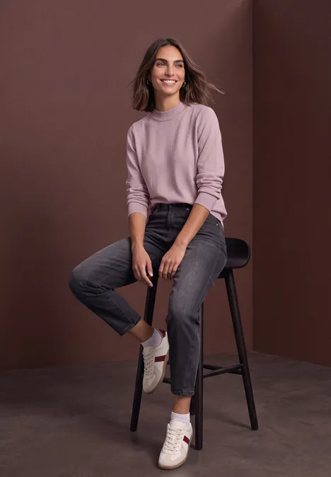 Pullover in Unifarbe mit Rundhals quiet rose Eine lächelnde Frau in rosafarbenem Pullover und dunkler Hose sitzt auf einem Barhocker vor braunem Hintergrund.