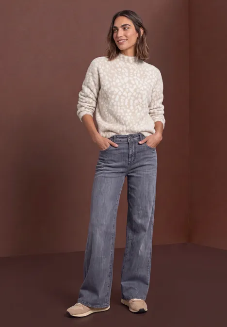 Frau im hellen Strickpullover und weiten Jeans vor braunem Hintergrund.