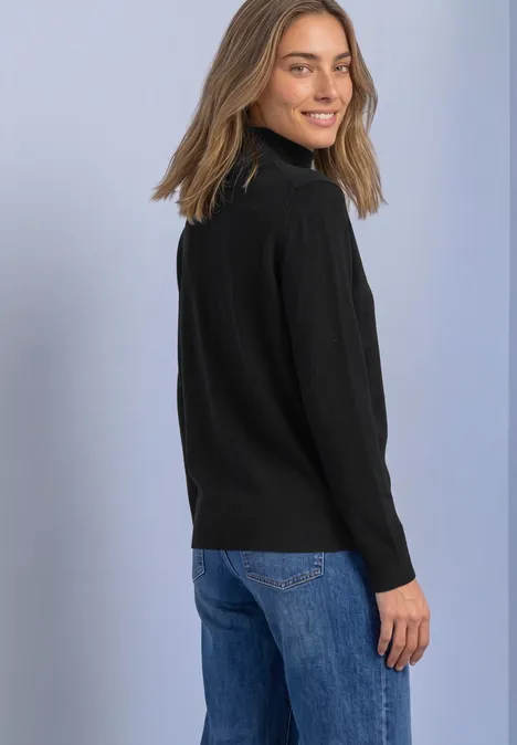 Junge Frau mit schwarzem Rollkragenpullover und blauen Jeans, schaut lächelnd über die Schulter vor hellem Hintergrund.