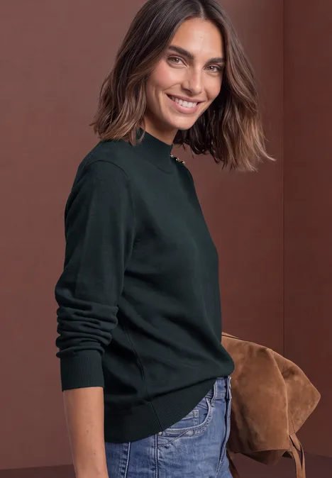 Lächelnde Frau in grünem Pullover und Jeans mit brauner Tasche vor rotem Hintergrund.