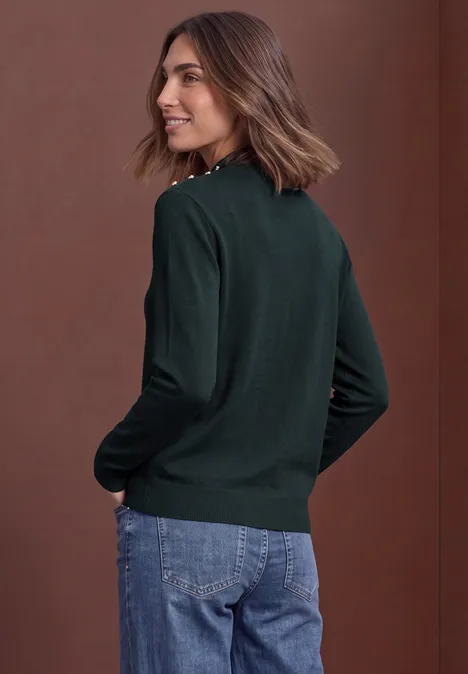 Frau im dunkelgrünen Pullover und Jeans, im Profil stehend, vor braunem Hintergrund.