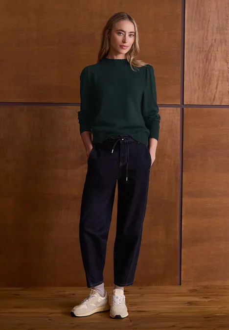 Langarm Pullover mit Turtle Neck und Raffung stormy green Frau steht vor Holzpaneelen, trägt grünen Pullover, dunkle Hose, helle Sneaker, Hände in den Taschen.