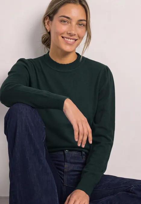 Langarm Pullover mit Turtle Neck und Raffung stormy green Frau sitzt lächelnd, trägt dunkelgrünen Pullover und blaue Jeans vor hellem Hintergrund.