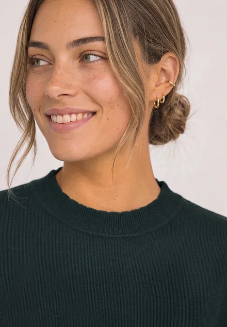 Langarm Pullover mit Turtle Neck und Raffung stormy green Frau mit braunem Dutt, mehreren goldenen Ohrringen und schwarzem Pullover, die leicht lächelt.