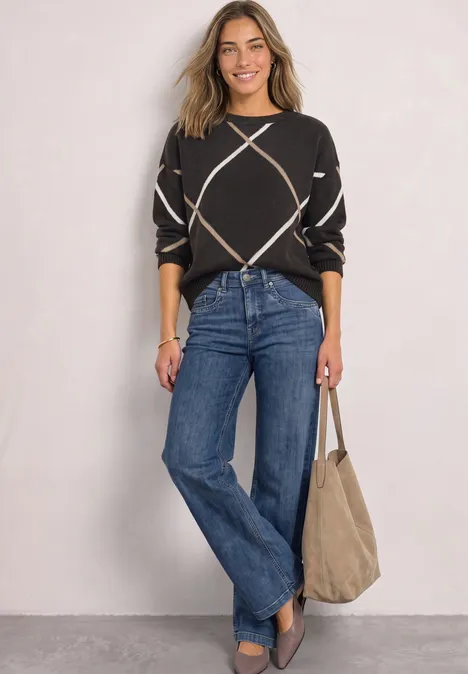 Frau in schwarzem Pullover mit Rautenmuster, blauen Jeans und beiger Tasche, lächelnd vor heller Wand.