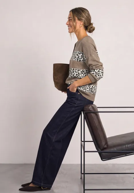 Sweter Jacquard z długim rękawem w stylistyce żakardowej z dekoltem w kształcie łódki sanded amber mel. Kobieta w wzorzystym swetrze i szerokich dżinsach opiera się o krzesło, trzyma kurtkę.