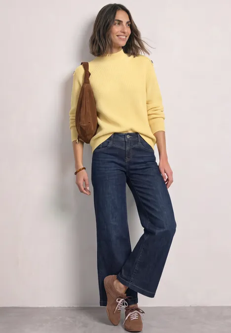 Frau lehnt an Wand, trägt gelben Pullover, weite Jeans, braune Schuhe und eine braune Tasche.