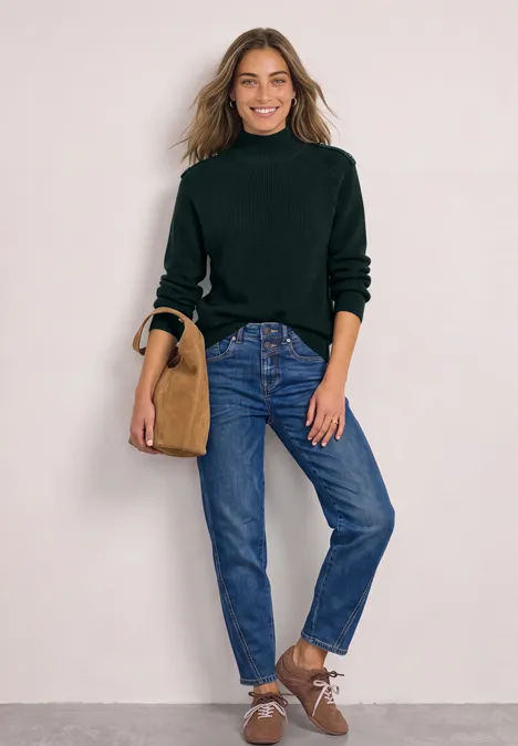 Frau in dunkelgrünem Rollkragenpullover, blauen Jeans und braunen Schuhen mit Tasche vor hellem Hintergrund.