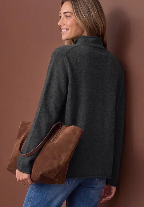Frau in grauer Jacke mit brauner Wildledertasche, lächelnd über die Schulter blickend.