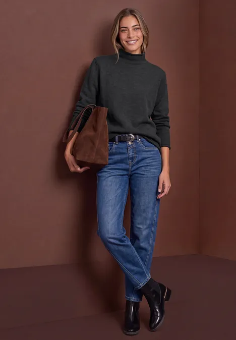 Eine Frau in Jeans, schwarzem Rollkragenpullover und Stiefeletten, trägt eine braune Tasche vor braunem Hintergrund.