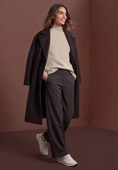 Frau in dunkelbraunem Mantel, beigem Pullover, weiter Hose und weißen Sneakers vor braunem Hintergrund.